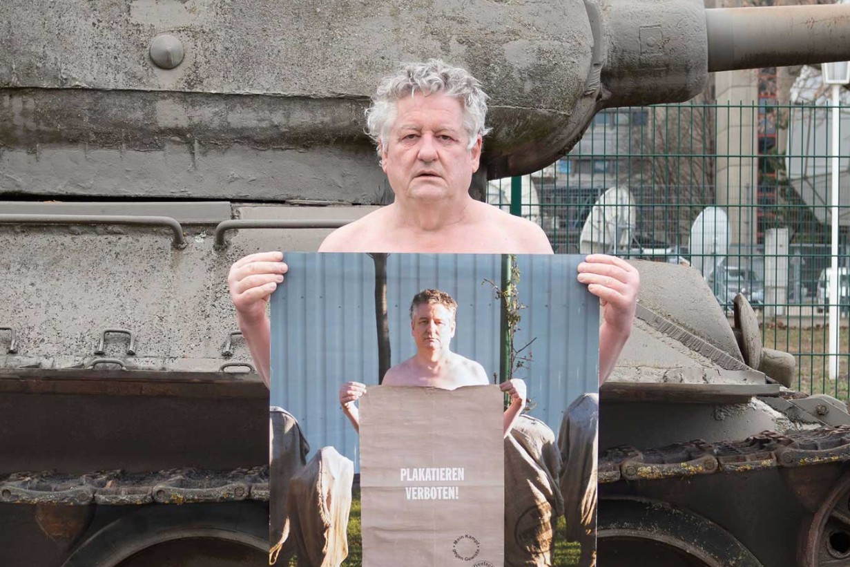 White man in front of a tank, he is holding a poster with a photo of himself on it in the same pose - holding a poster.