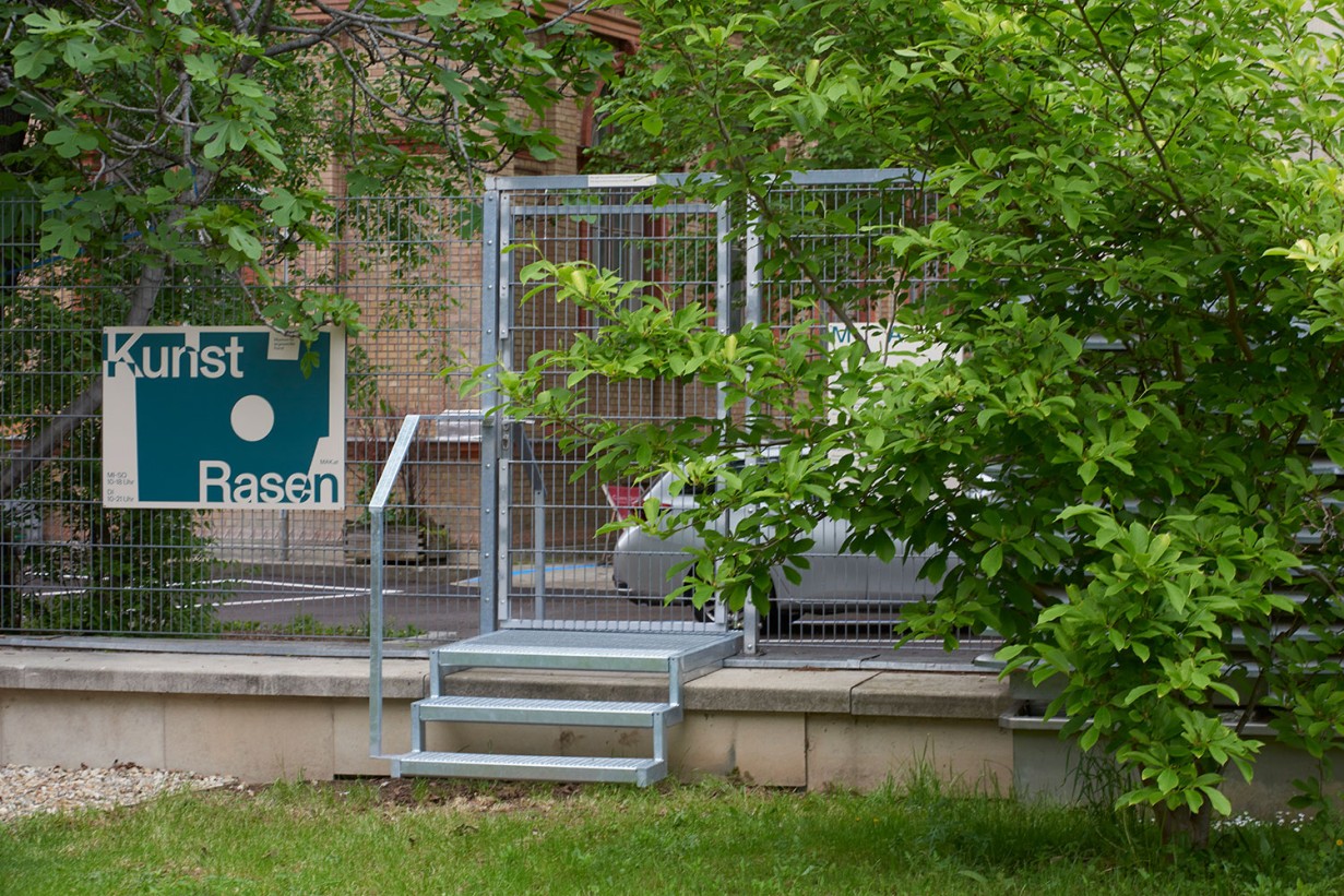 A lattice fence with gate, green tree and a poster on the fence, on it to read "KUNSTRASEN".