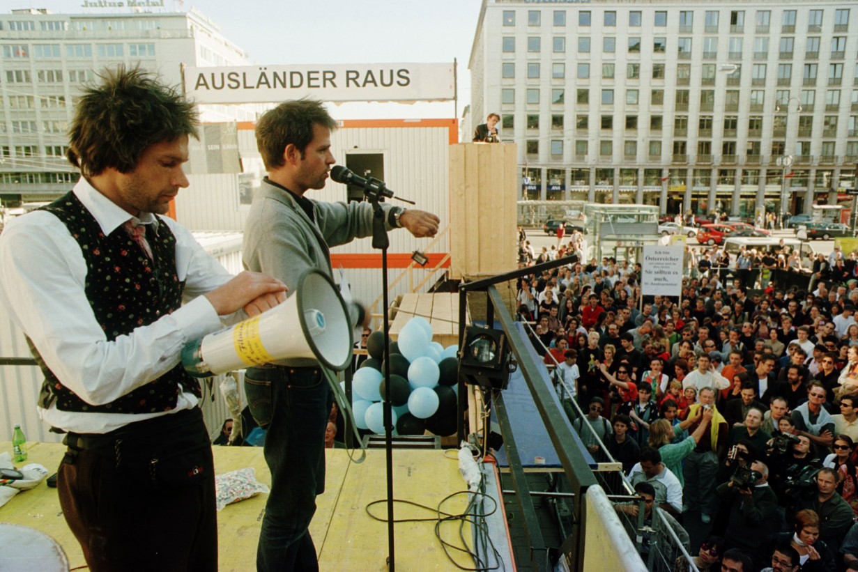 Christoph Schlingensief in front of containers talking to crowd (Bitte liebt Österreich)