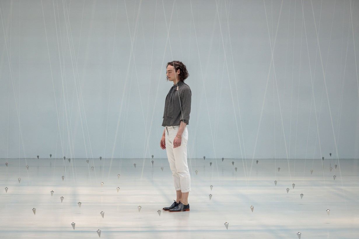 Eine Ausstellungshalle mit Pendeln, die an Schnüren von der Decke hängen. 1 Person bewegt sich zwischen den Schnüren.