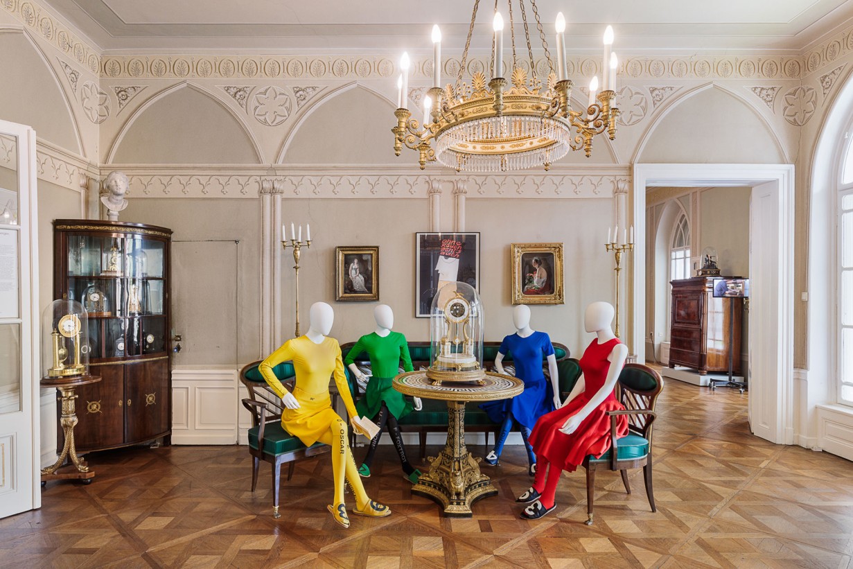 4 Schaufensterpuppen sitzen gekleidet in der Kollektion Fashion is Fast von Anna-Sophie Berger um einen Tisch. 