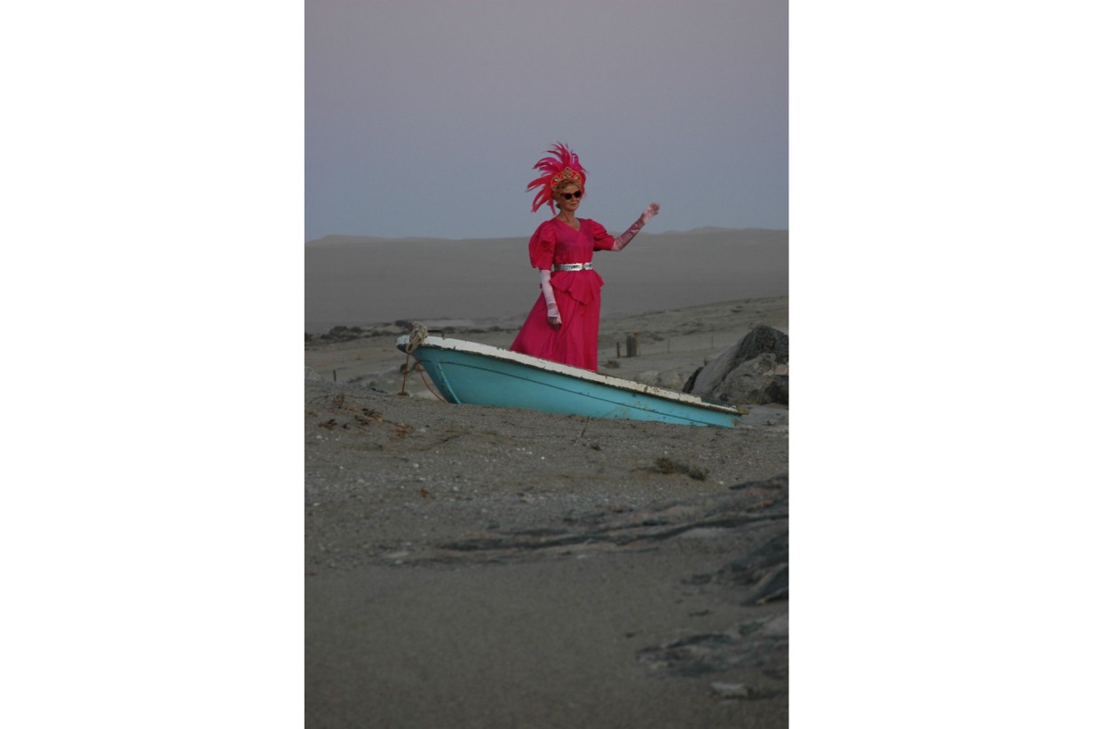A person wearing a pink dress and feathers on their head is standing in a boat in the desert