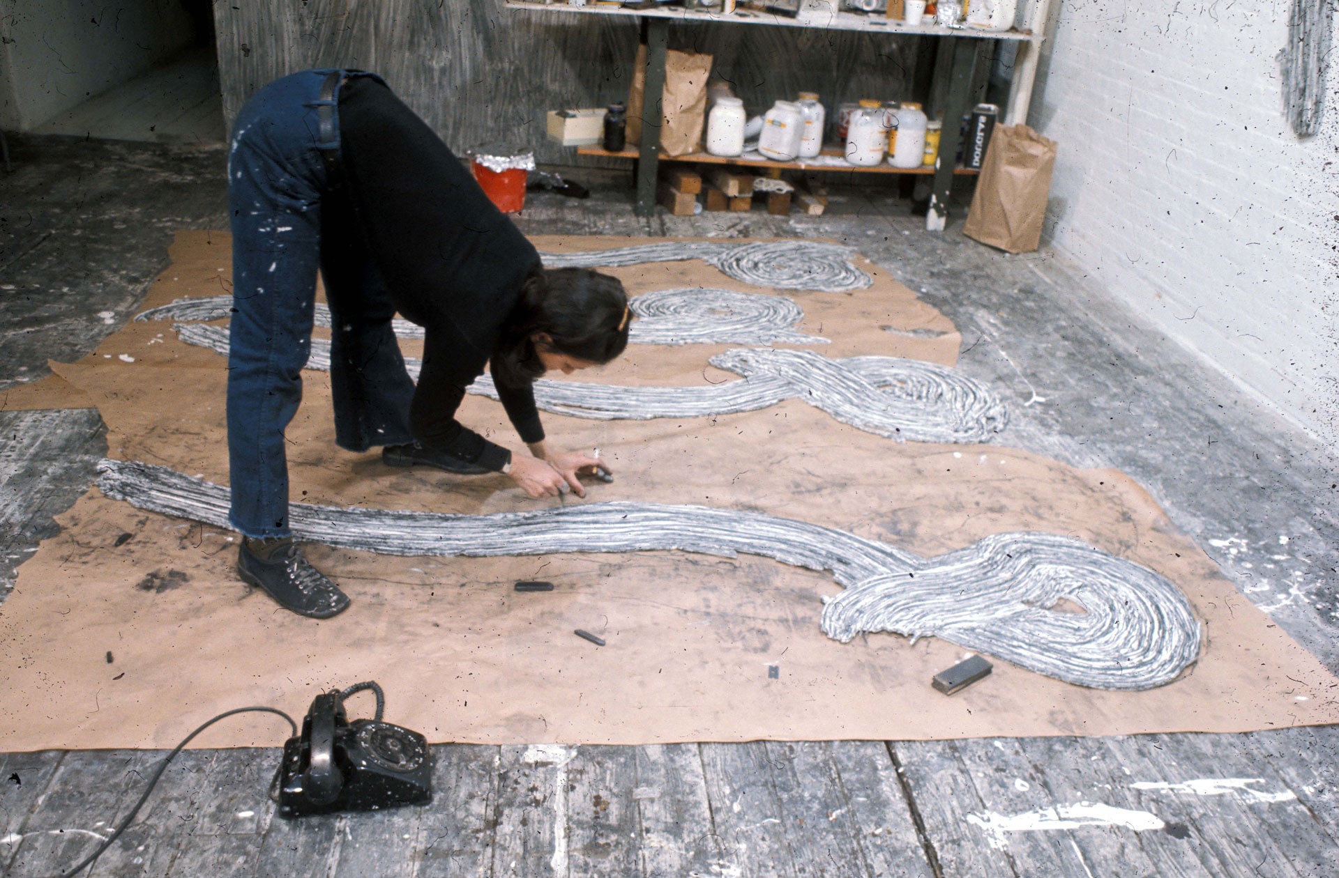 A woman bends over to continue working on a sculptural work of art on the floor. In one hand she holds a cigarette.