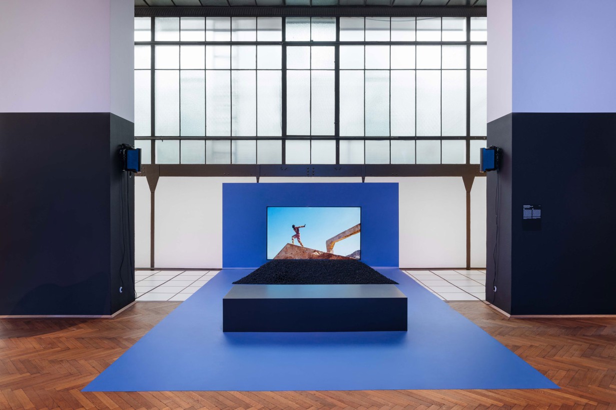 Exhibition hall, black pedestal, behind it a video, blue floor 
