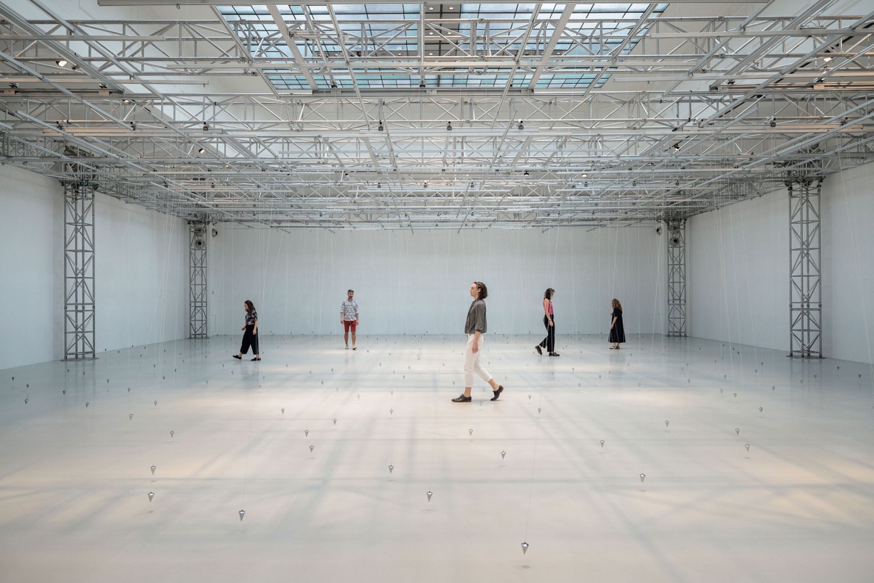 Eine Ausstellungshalle mit Pendeln, die an Schnüren von der Decke hängen. 5 Personen bewegen sich zwischen den Schnüren.