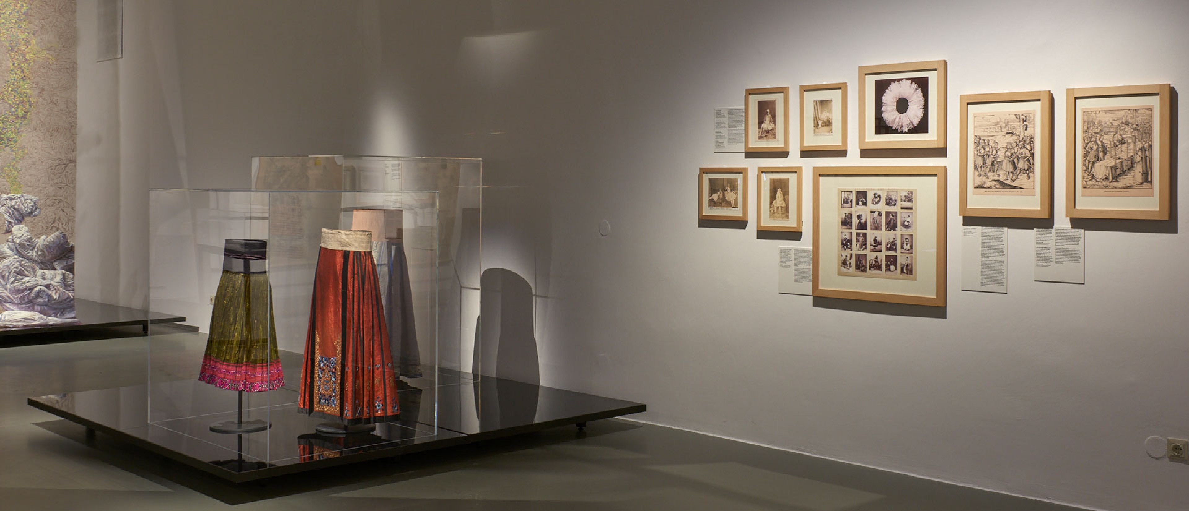 Ausstellungsraum mit Objekten: Faltenröcke, Malerei, Fotografien an den Wänden.