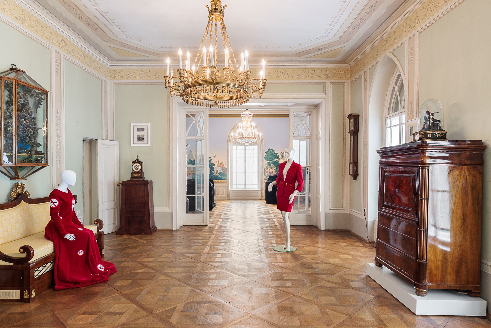 Two mannequins are in a room in the Geymüllerschlössel, one is sitting on a canapé, the other is standing. Both are wearing red dresses
