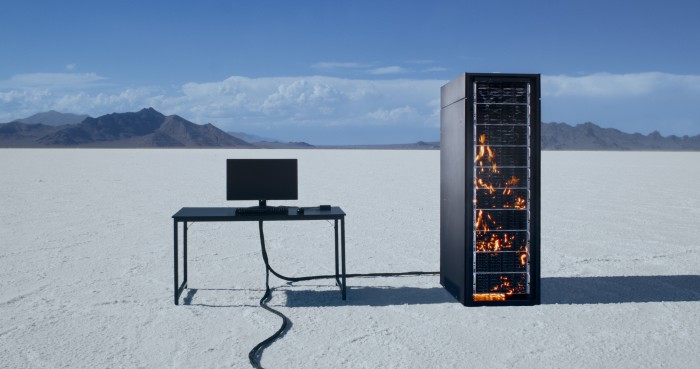 Ein Computerbildschrim auf einem Schreibtisch, daneben ein Servertower in Flammen in einer Wüstenlandschaft.