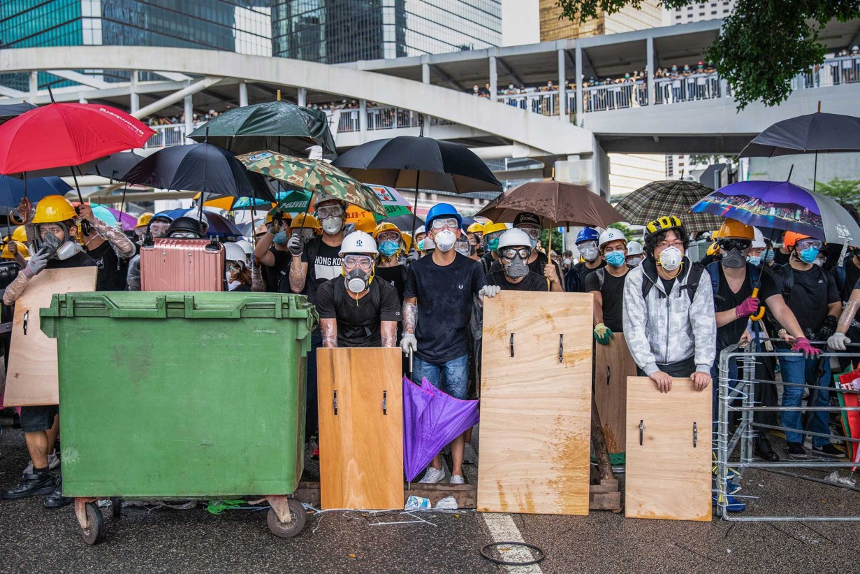 Aktivist*innen mit Masken, Helmen, selbst gebauten Schilden und Regenschirmen, um sich vor den Tränengas- und Wasserwerfereinsätzen der Polizei zu schützen. Im Gegensatz zu den ortsbezogenen Strategien von 2014 waren die Proteste von 2019–2020 von „fluiden“ Taktiken geprägt, die unter dem Motto „Be Water“ standen. 