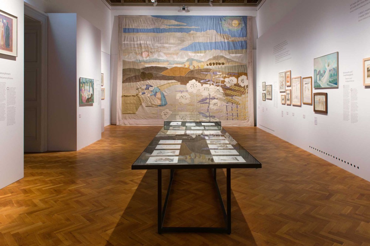 Ausstellungsraum, in der Mitte eine Vitrine, an den Wänden gerahmte Bilder, frontal ein Wandteppich