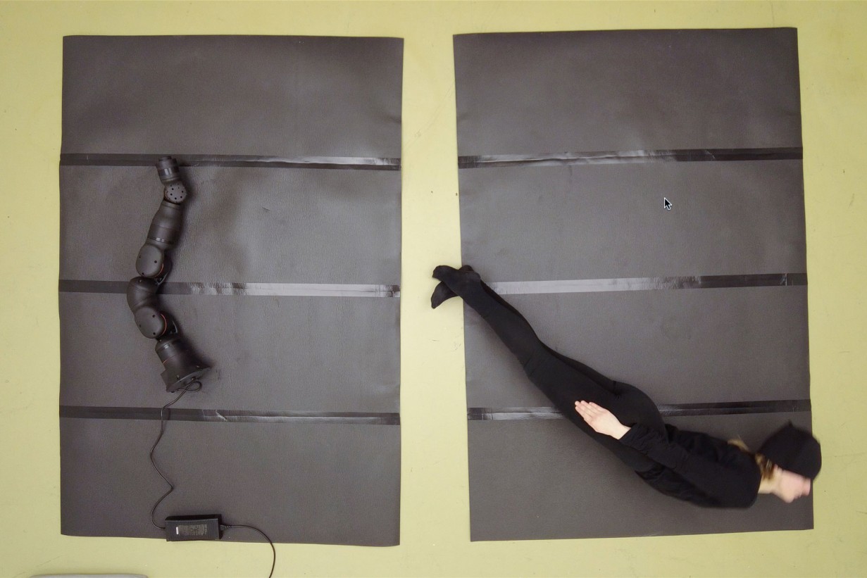 A dancer is lying on the floor and is being photographed from a bird's-eye view; a robotic arm is lying next to him