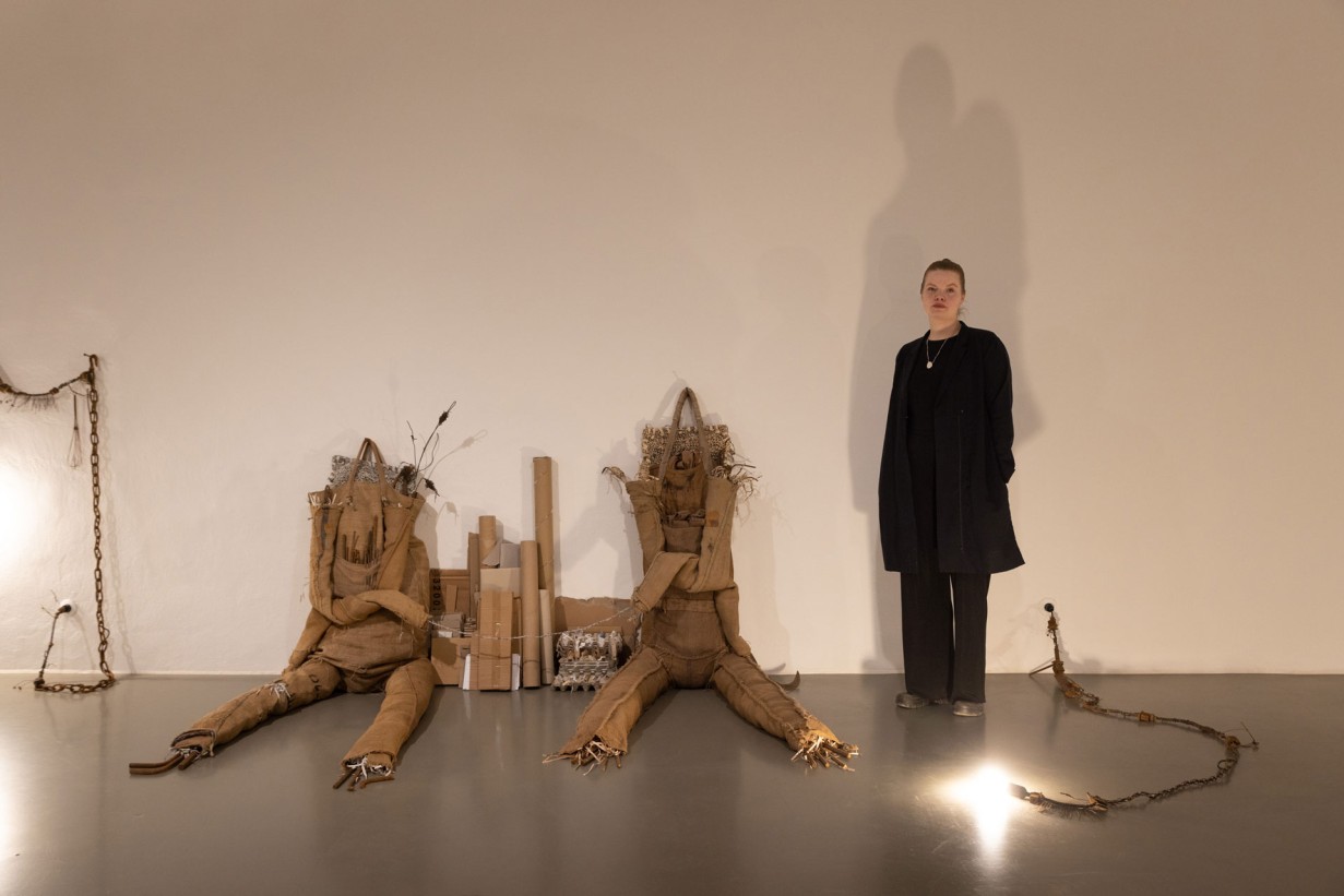 Artist Birke Gorm in the exhibition space, two scupltures made of jute bags leaning against a wall. 