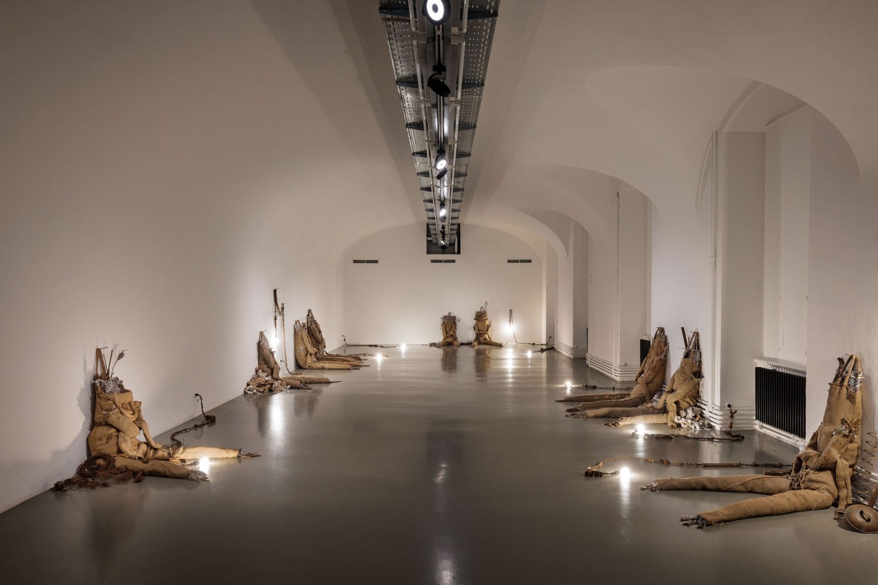 Space with scupltures made of jute bags, leaning against the wall. Accompanying the figures are small sculptures made of scrap metal and electrical wiring, with attached bulbs serving to light the exhibition space.