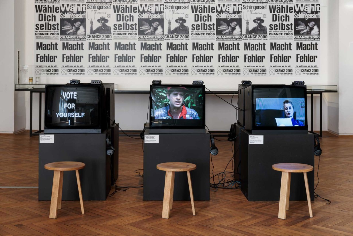 Plakatwand und Fernseher mit Slogans "Macht Fehler", "Wähle dich selbst", Wählt Schlingensief"