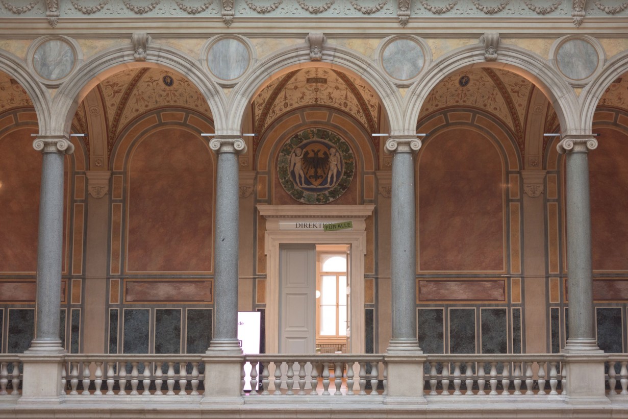 Ein historisches Gebäude mit Arakden und Säulen, dahinter eine Tür beschriftet mit "Direktion für alle"