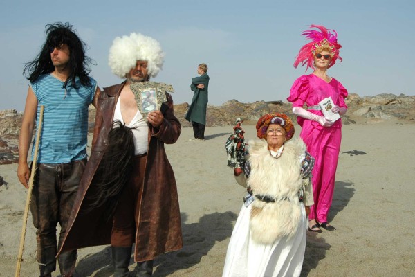 Five people wearing wigs stand in a desert-like setting.