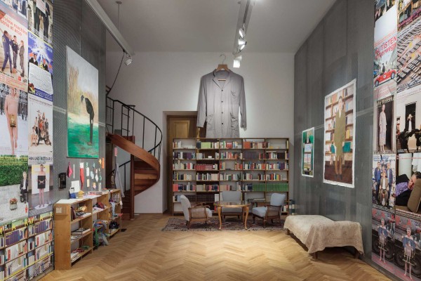 Central view of the exhibition space, left and right are many posters on the wallsr, frontal a kind of living room (library of unread books). An oversized coat hangs from the ceiling. 