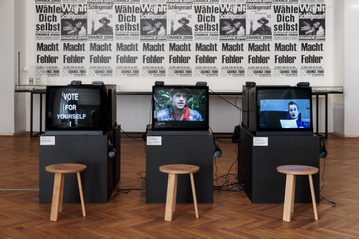 Plakatwand und Fernseher mit Slogans "Macht Fehler", "Wähle dich selbst", Wählt Schlingensief"