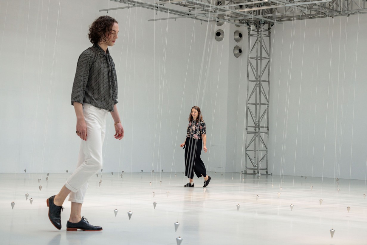 Eine Ausstellungshalle mit Pendeln, die an Schnüren von der Decke hängen. 2 Personen bewegen sich zwischen den Schnüren.