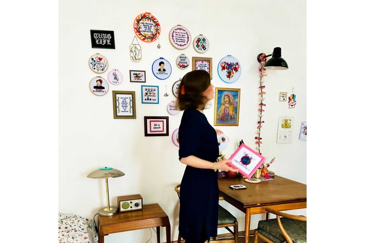 A woman stands in a room holding a picture with a frame. Many cross-stitch objects on the wall.