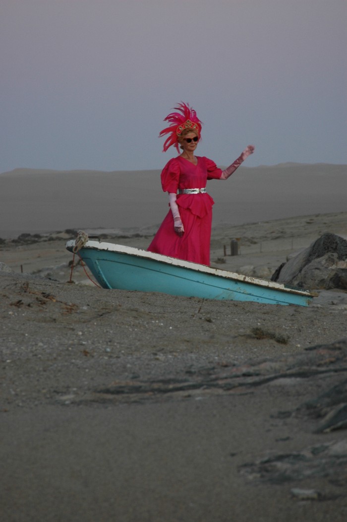 Person in pinken Kleid und mit Federn am Kopf steht in einem Boot in der Wüste